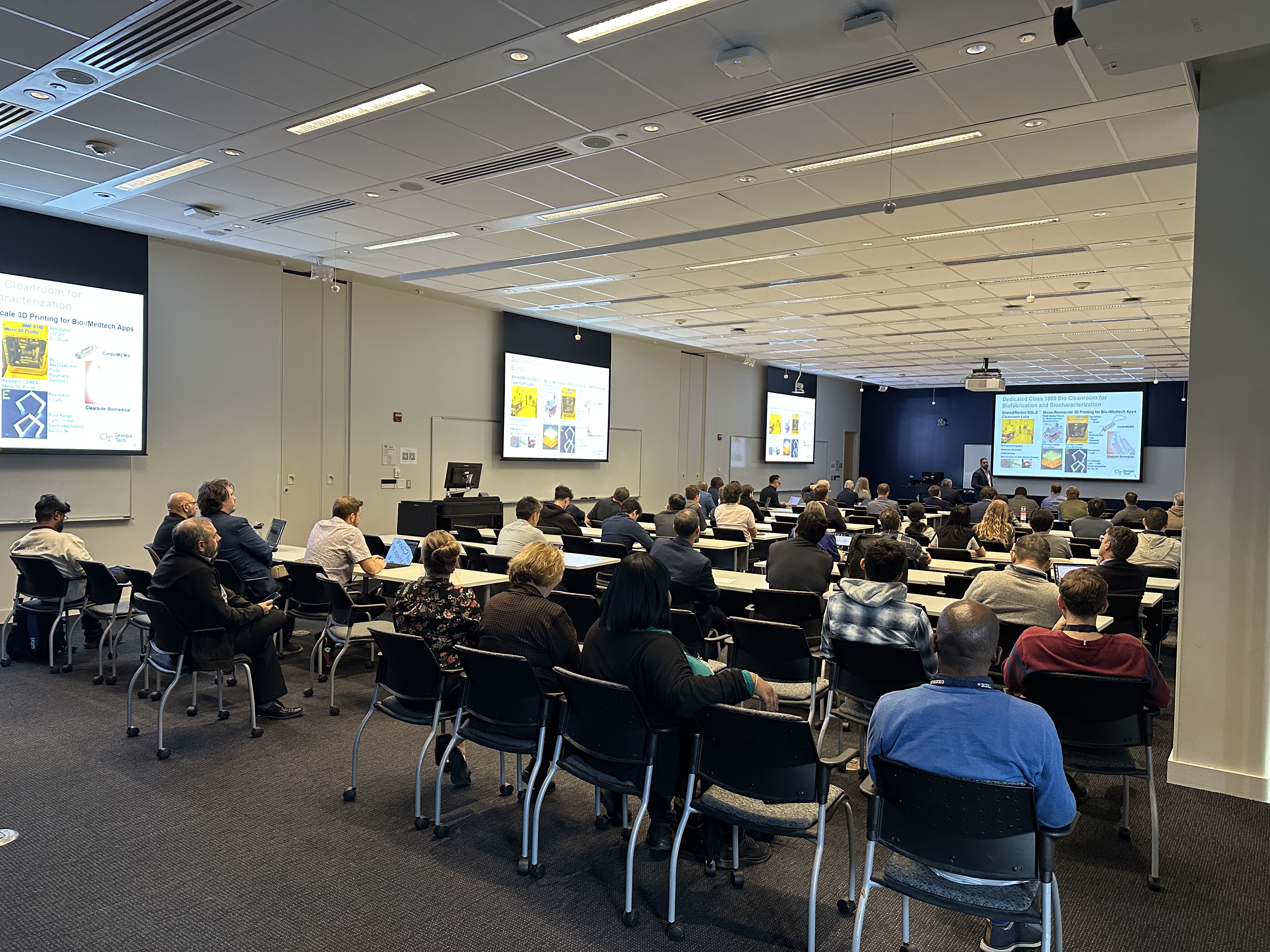 Attendees listen to a research presentation in a conference room while slides on advanced microelectronics and nanotechnology topics are displayed on large screens.