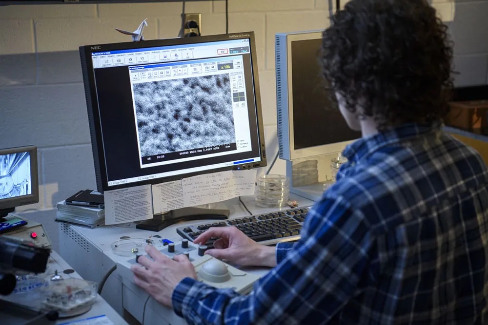A student researcher operates a scanning electron microscope workstation, analyzing high-resolution images of nanomaterials and microstructures displayed on a computer monitor.