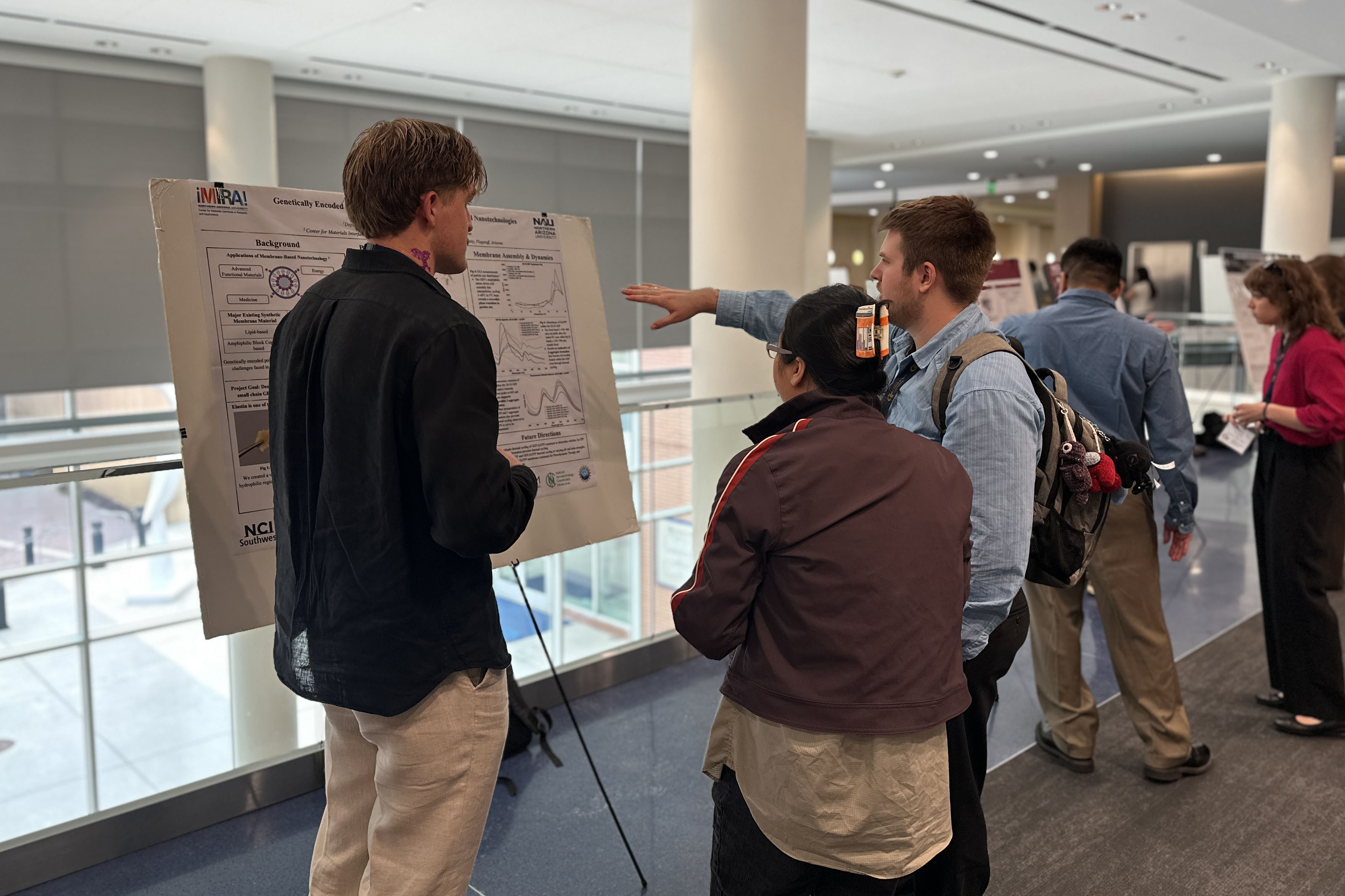 Researchers and conference attendees discuss a scientific poster presentation during a research symposium, examining data, charts, and findings displayed on a poster board in an academic conference setting.