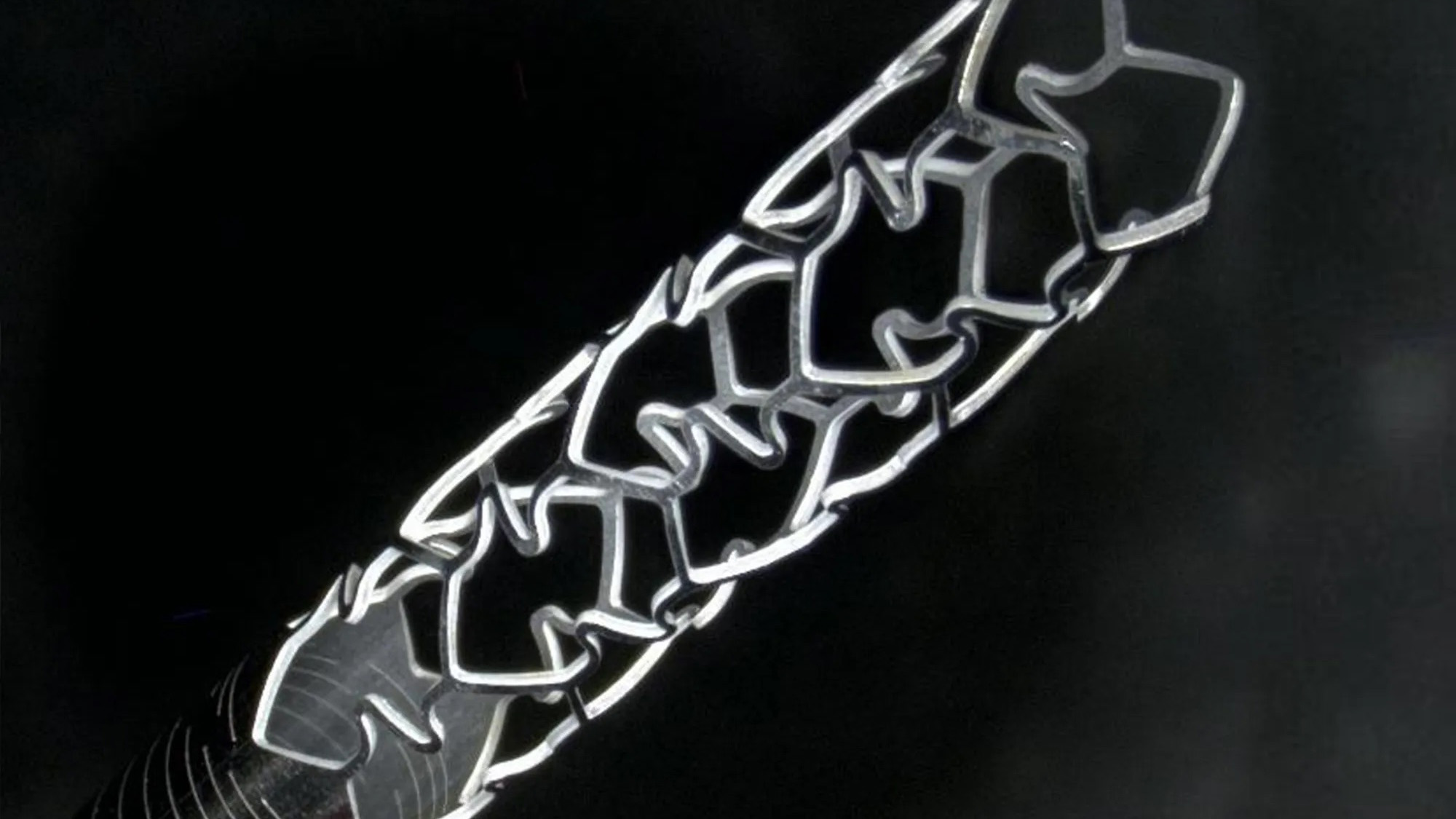 Scanning electron microscope (SEM) images show a textured titanium alloy surface with layered microscale patterns created by laser irradiation, including a magnified inset highlighting fine surface features and scale measurements.  Close-up of a metal cardiovascular stent with an intricate lattice structure mounted on a catheter against a dark background.