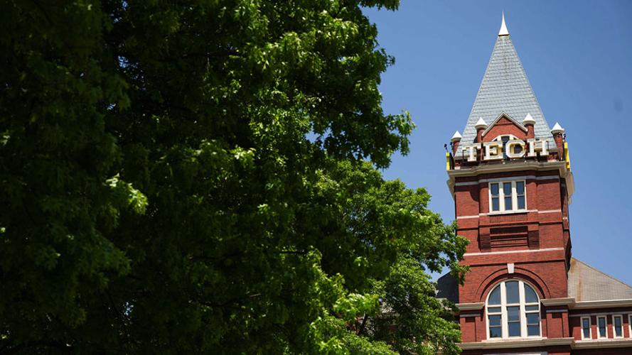 Georgia Tech’s historic Tech Tower rises above leafy green trees on a clear day, featuring a red brick facade, arched windows, and a pointed gray roof with the word “TECH” displayed prominently near the top.